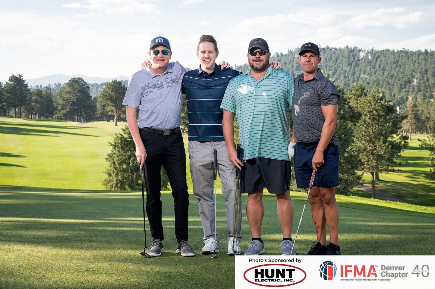 Four men on a golf course pose with clubs. One has arm around another. Green grass, trees, and blue sky in background.