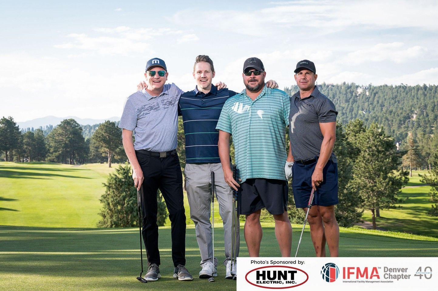 Four men posing together on a golf course. They wear golf attire and hold clubs, with a green landscape backdrop.