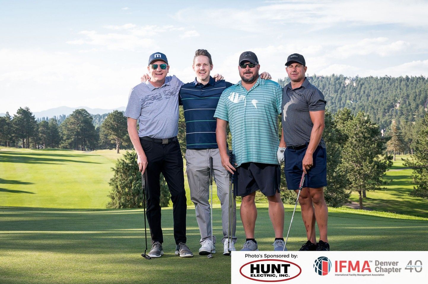 Four men in golf attire pose on a course. Mountain backdrop, sunny day.