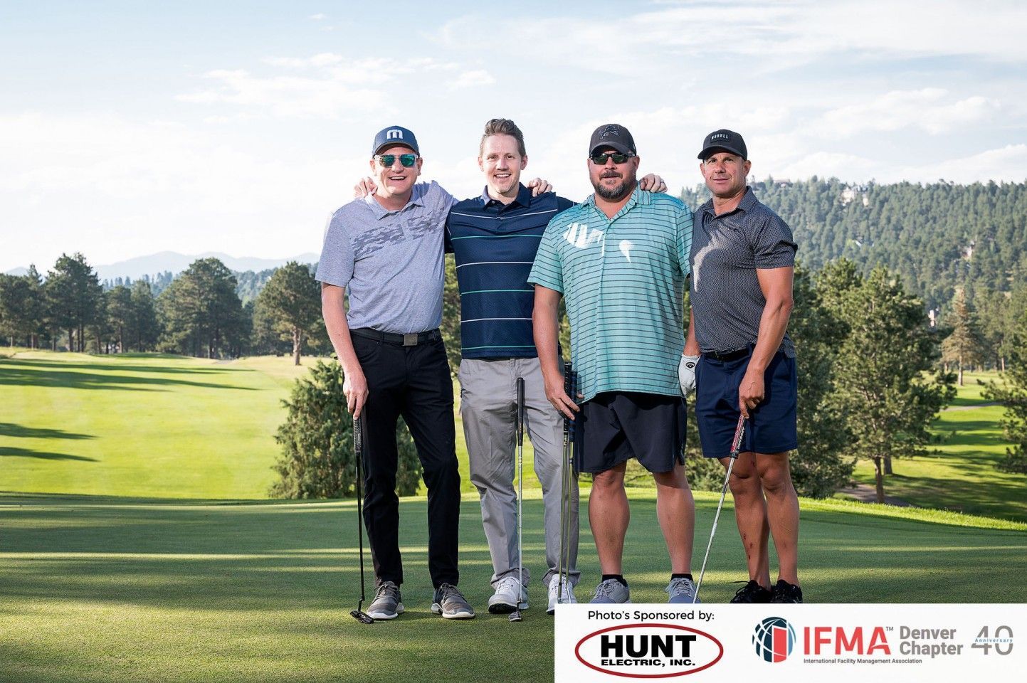 Four men pose on a golf course. They wear golf attire and hold clubs, with a forested mountain backdrop.