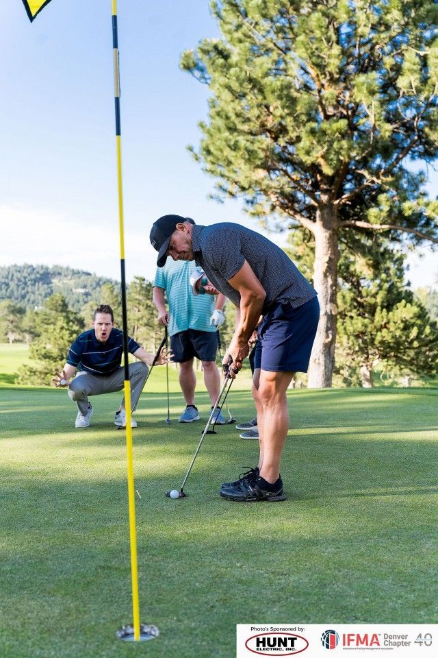 Man putting golf ball on a green, two others watch. Sunny day.