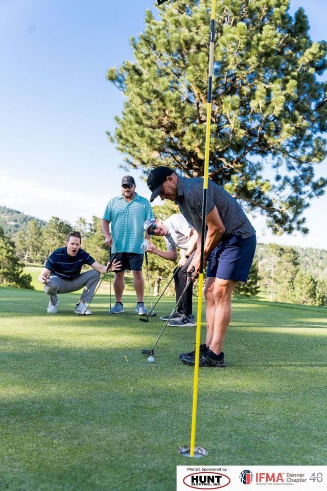 Golfers practicing putting on a green; one lines up a putt while others watch, a yellow guide post in foreground.