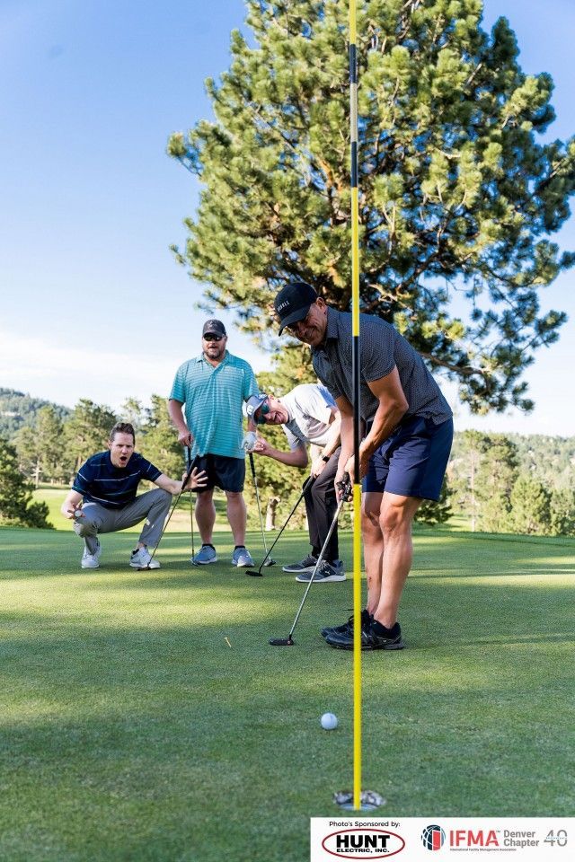 Golfers putting on a green, one using a straight-line aid. Others watch. Sunny day, golf course setting.