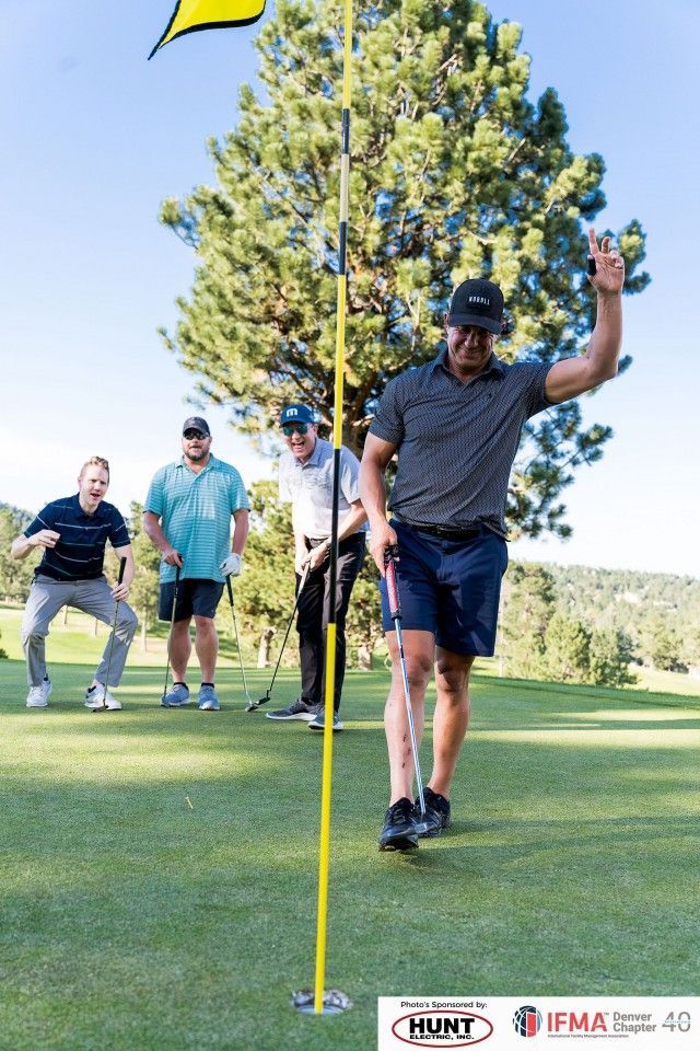 Golfers celebrate on the green; one waves his arm, flag in view, other players look on a sunny day.