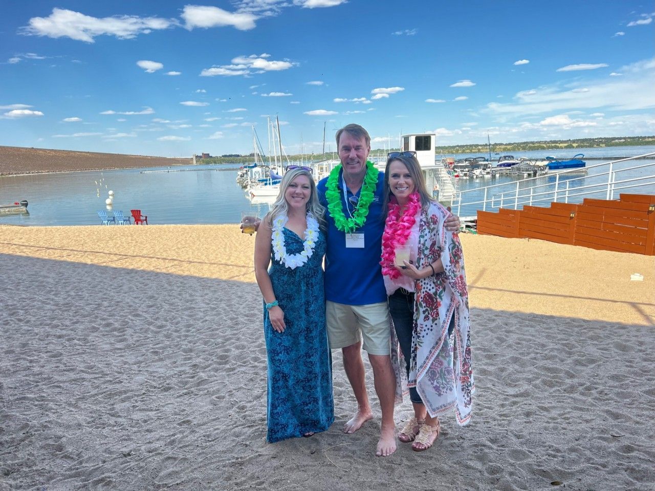 Three people on a sandy beach wearing leis, posing near water and boats.