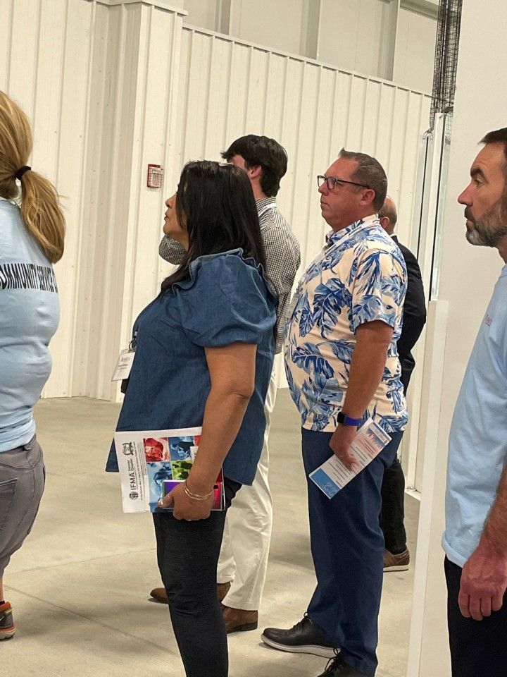 People standing in a line indoors, some holding papers. One man wears a blue floral shirt.