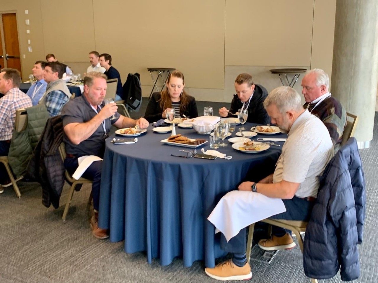 People eating at a round table, possibly at a business meeting. Blue tablecloth. Other people in the background.