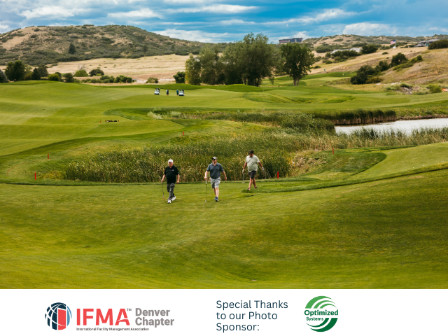 Three men walking on a green golf course, a pond, and hills in the background. Cloudy sky overhead.