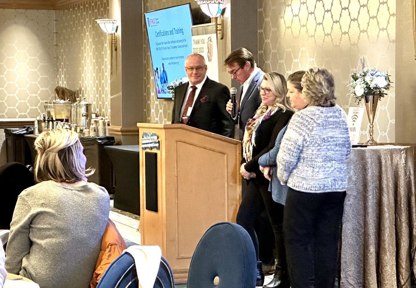 A man speaking at a podium with others standing beside him at an event.