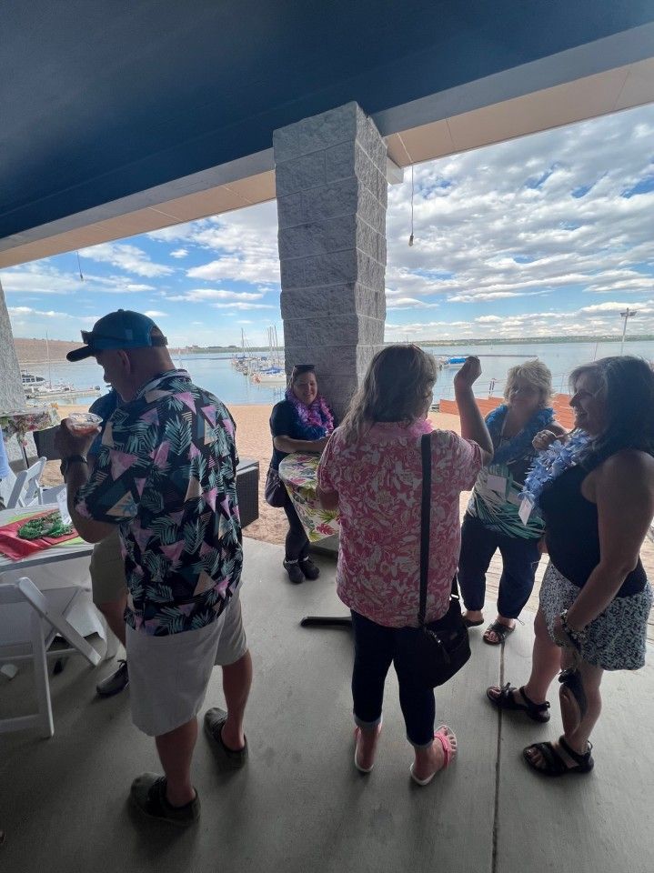 Group of people under a covered area, looking out at a body of water. Some are conversing, others viewing a table.