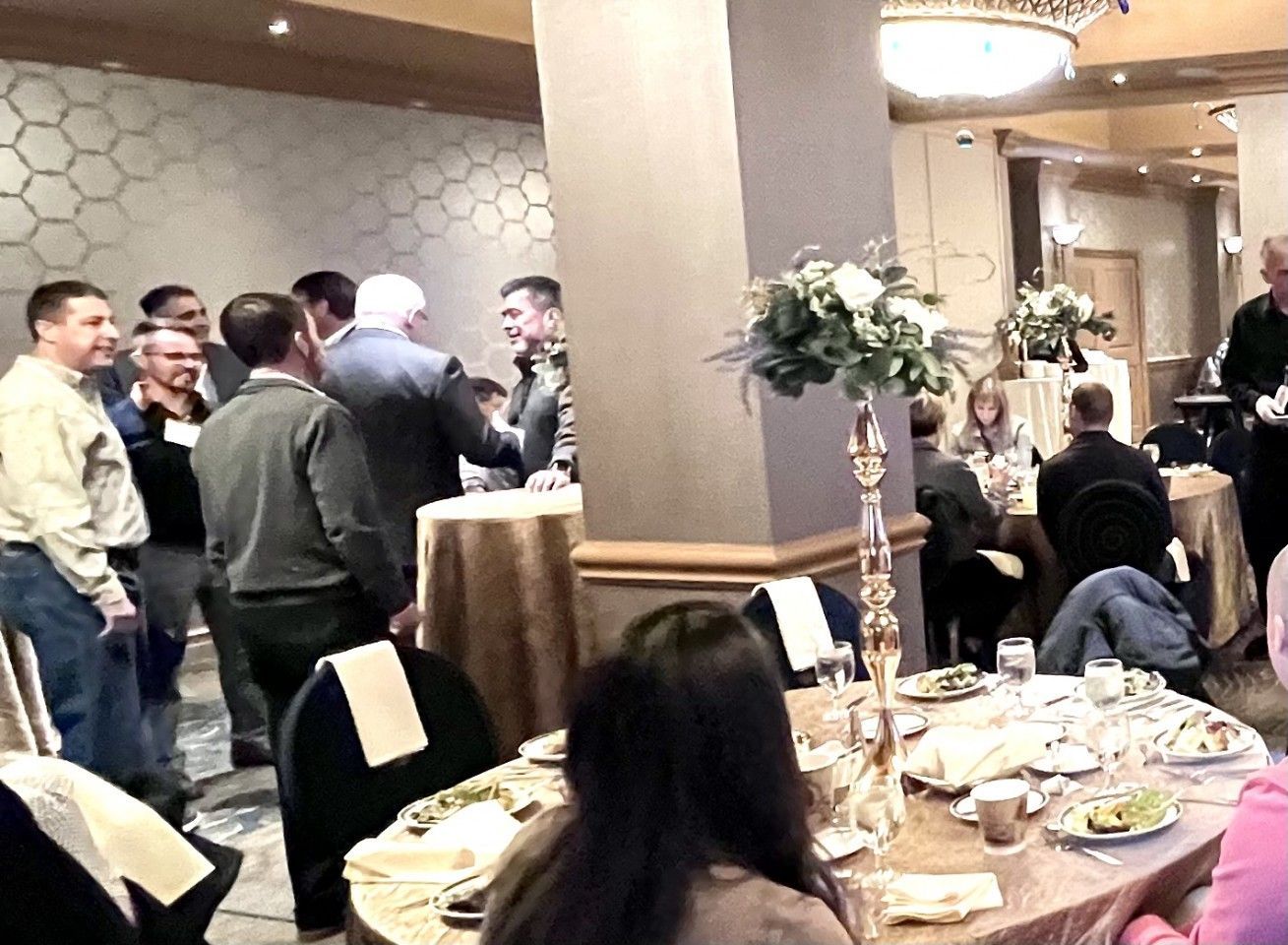 People at a gathering with tables, floral arrangements, and a group conversing by a pillar in a well-lit room.