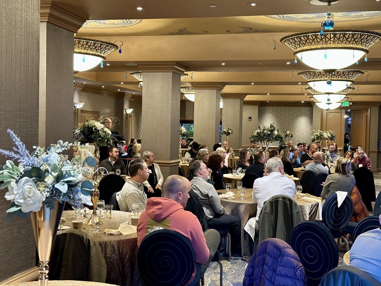 Large group of people seated at tables in a decorated ballroom for an event; floral centerpieces and ornate lighting.