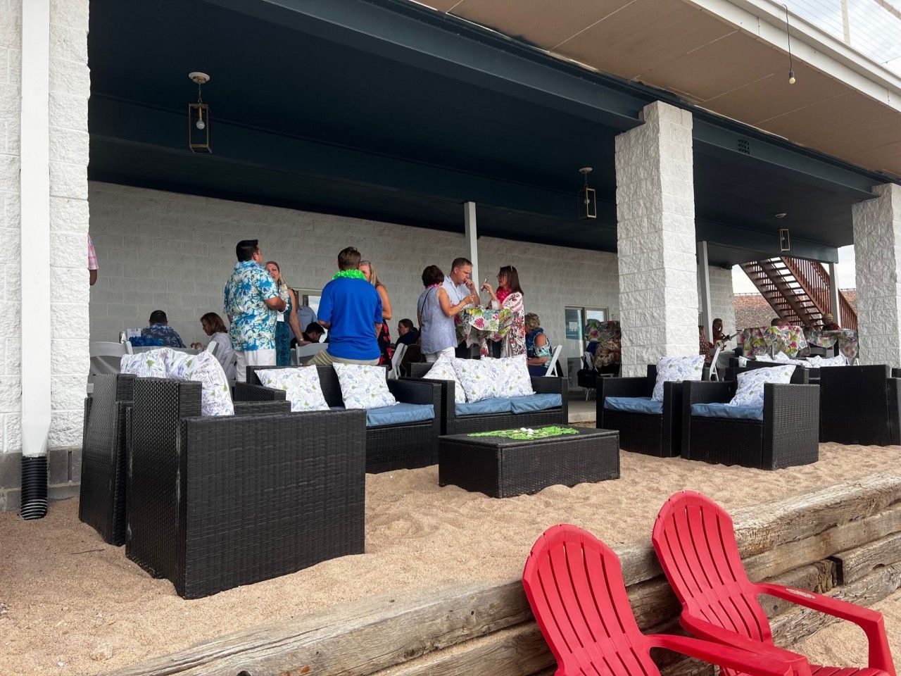 Outdoor event under a canopy with people mingling. Patio furniture, red chairs, and sandy ground.