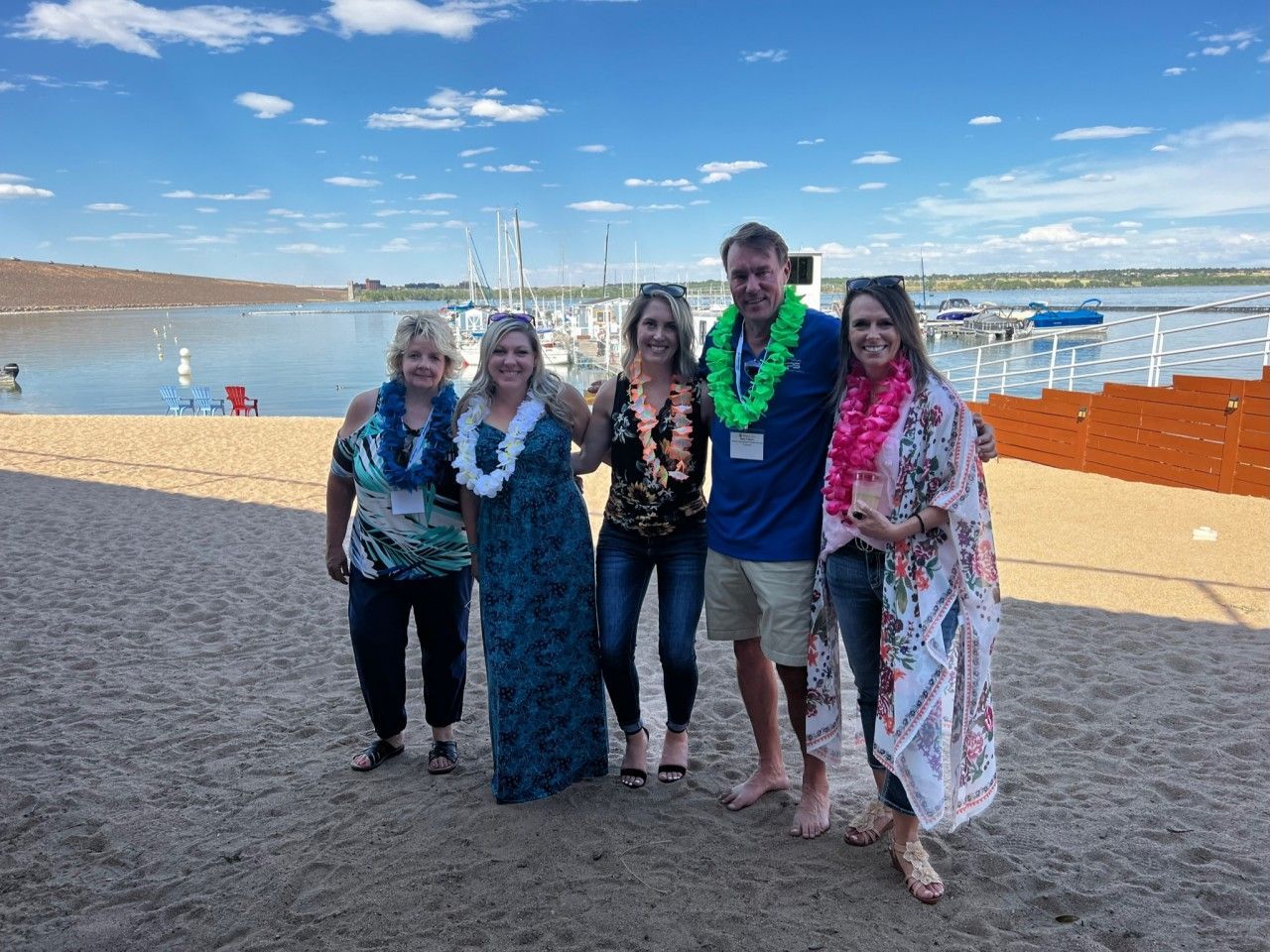 Five people at a beach, wearing leis. They are smiling, with a lake and boats in the background.