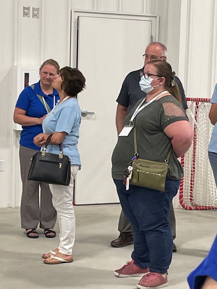 Four people stand in a white room. Two women in light blue tops converse, while a third woman in green and a man look on.