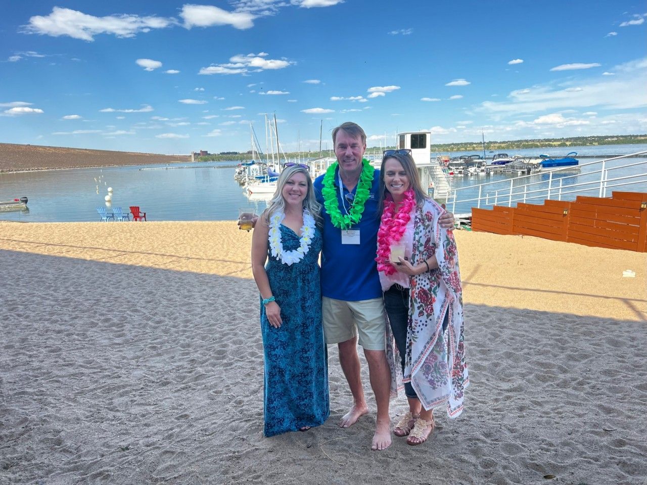 Three people on a sandy beach wearing leis, smiling at the camera. Boats and blue water are in the background.