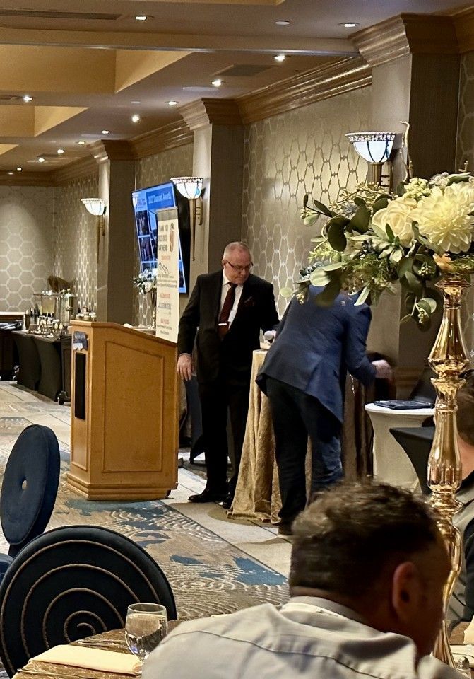 Man in suit at podium; another person near table with floral arrangement; event setting.