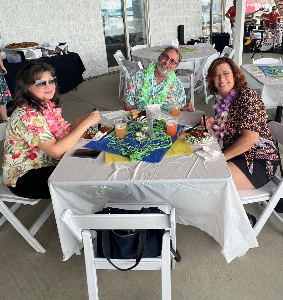 Three people at a table, wearing leis and Hawaiian shirts, enjoying a meal outdoors.