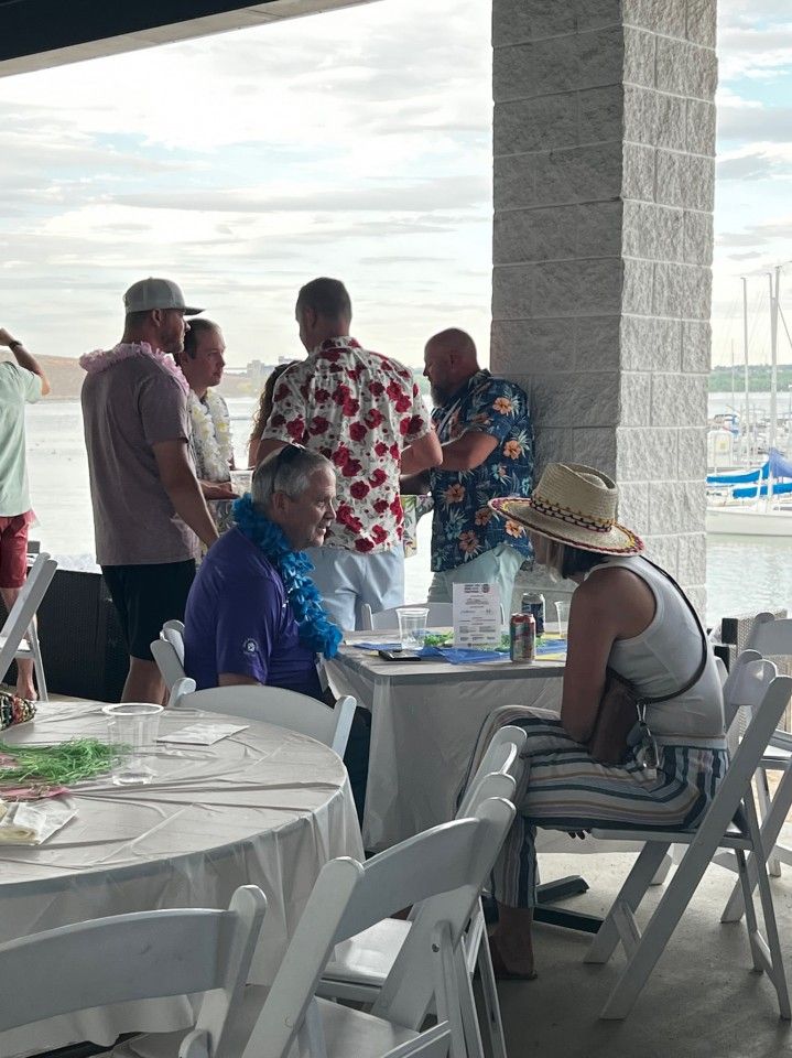 People in Hawaiian shirts at a waterfront event. A woman sits, others socialize near a table under a pavilion.