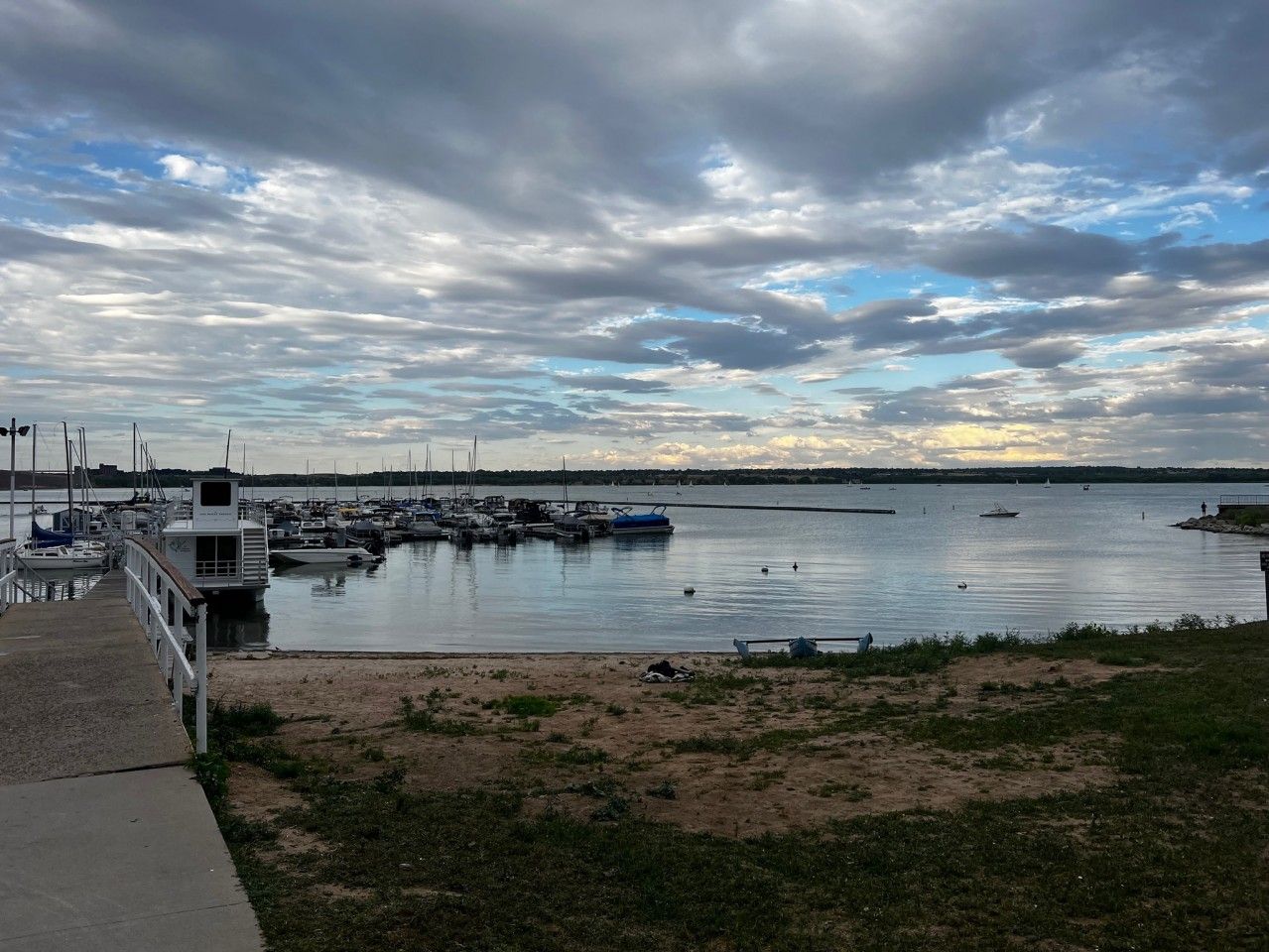 Boats docked in harbor under cloudy sky at a marina, with a sandy beach in the foreground.