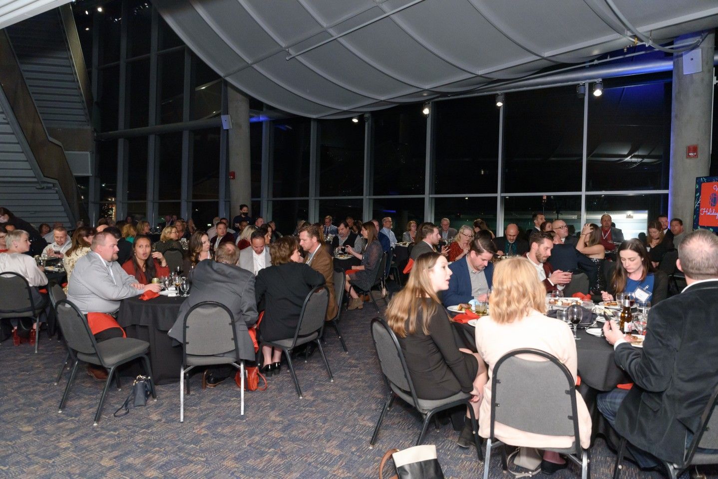 Large group of people seated at round tables in a modern venue, likely an event or reception.