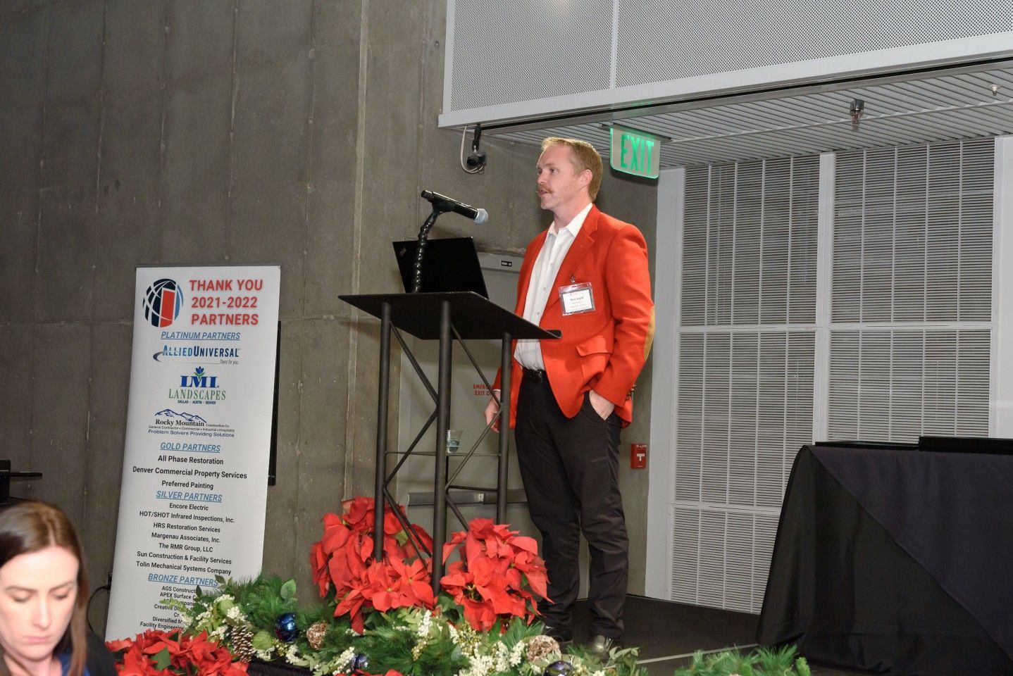Man in orange jacket speaks at a podium. Banner beside him, poinsettias in foreground.