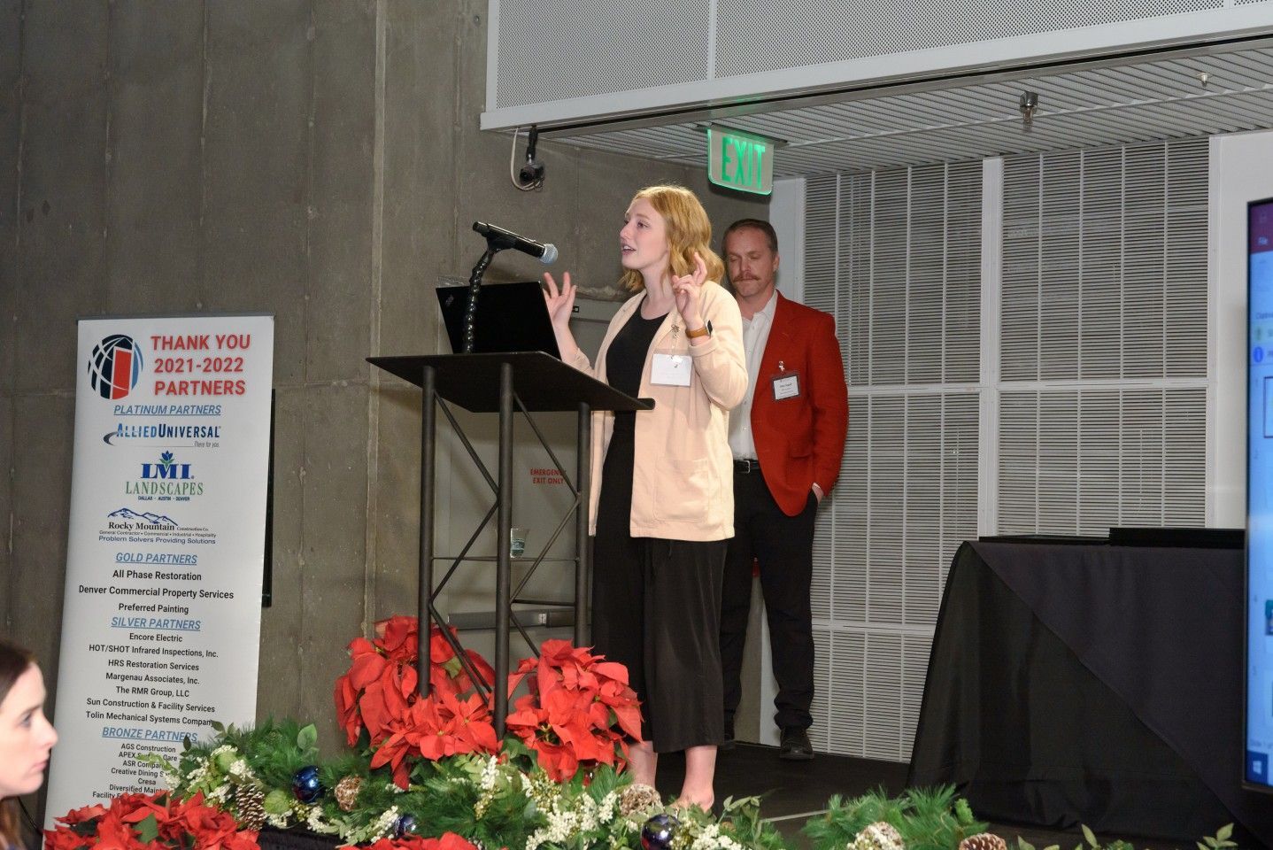 Woman speaking at a podium, man stands behind. Stage decorated, banner on left.
