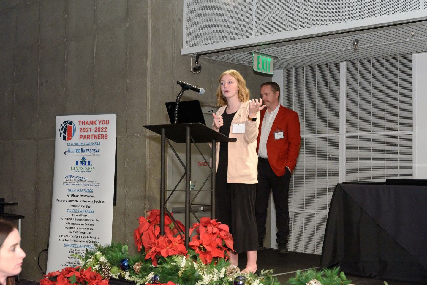 Woman speaking at podium, another person behind her. Event with holiday decorations.