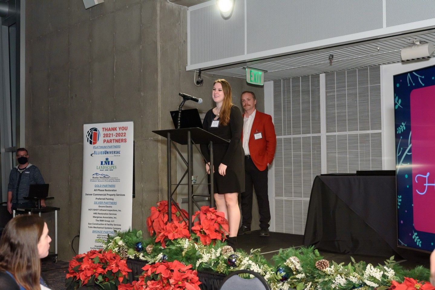 Woman speaking at podium, man stands behind. Stage decorated with poinsettias and greenery.