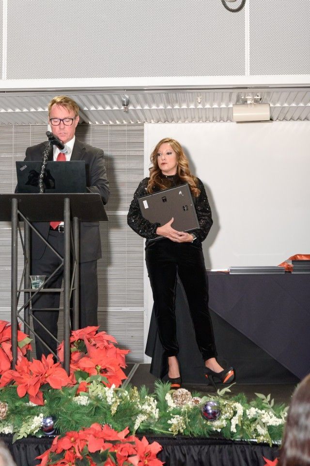 Man at podium, woman holding a presentation folder, standing on a stage decorated with poinsettias.