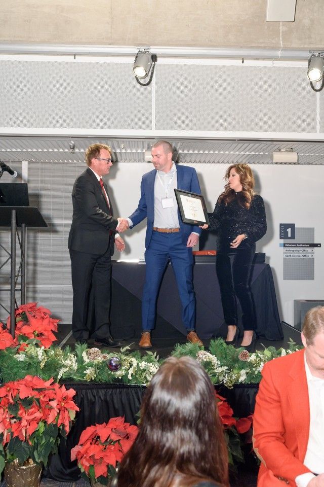 Man in suit shakes hands, another receives a document from a woman; stage with poinsettias.