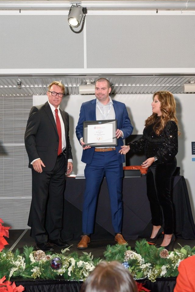 Man in blue suit holding an award on stage, flanked by two people, holiday decorations.