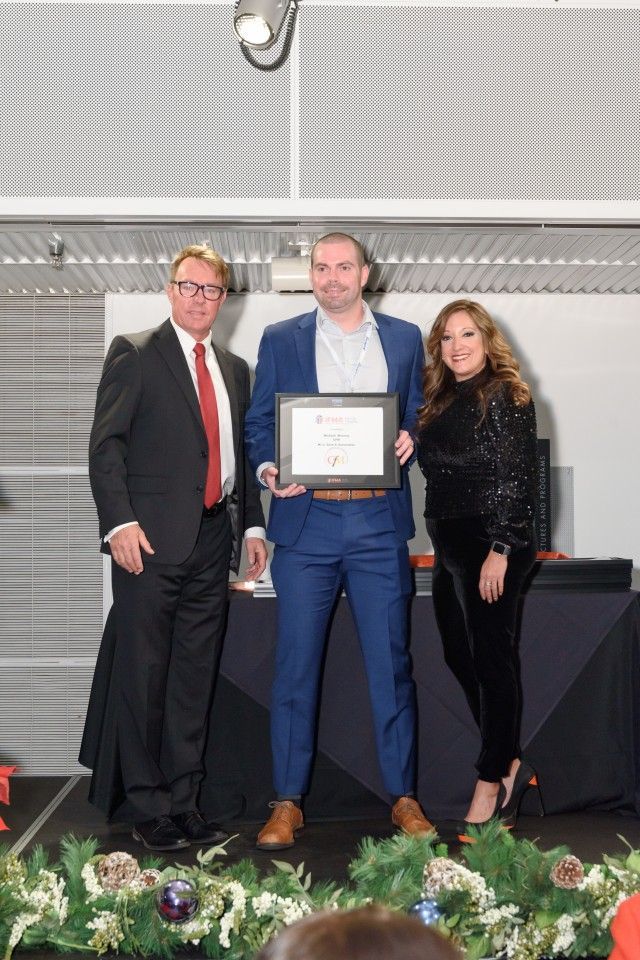 Man in suit holding award, flanked by two people, on stage with decorations.