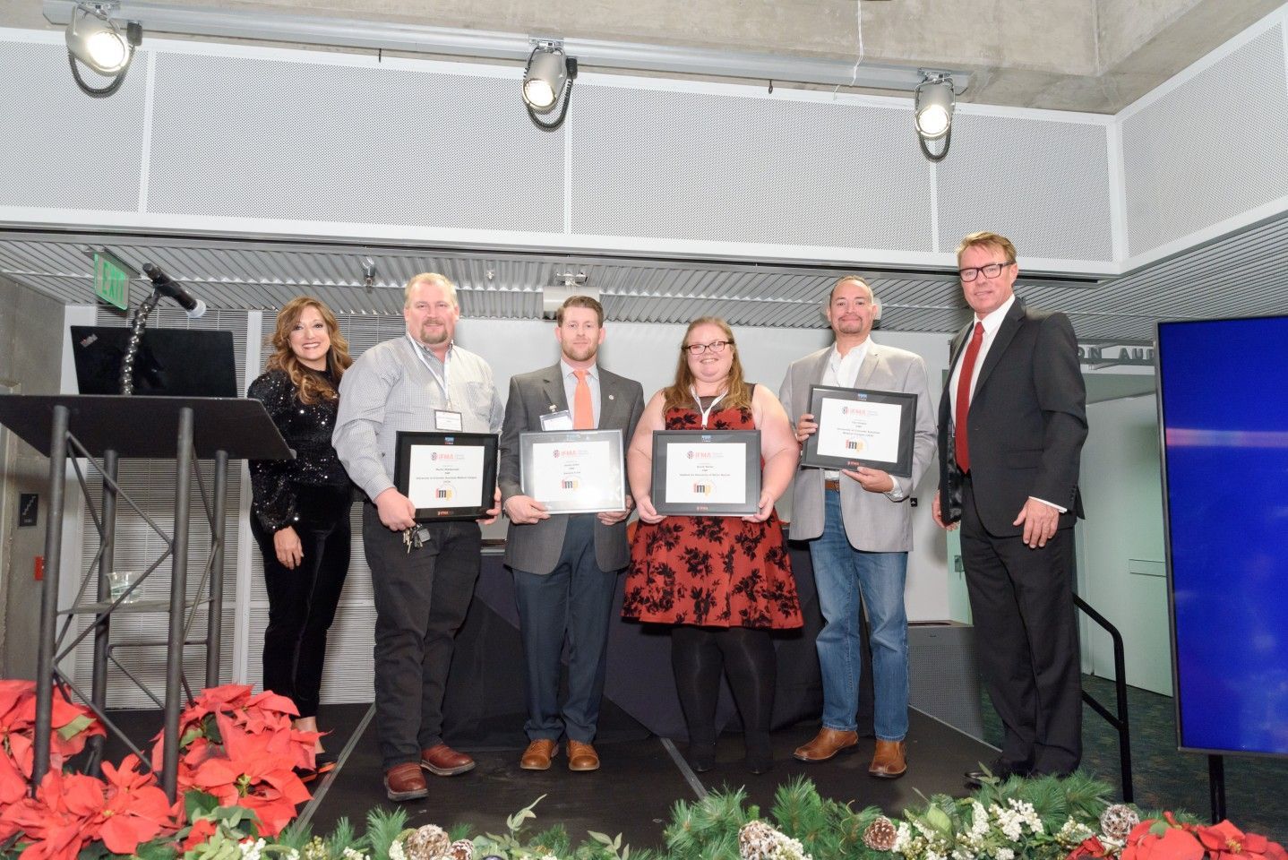 Group of people holding framed certificates on a stage, with holiday decor.