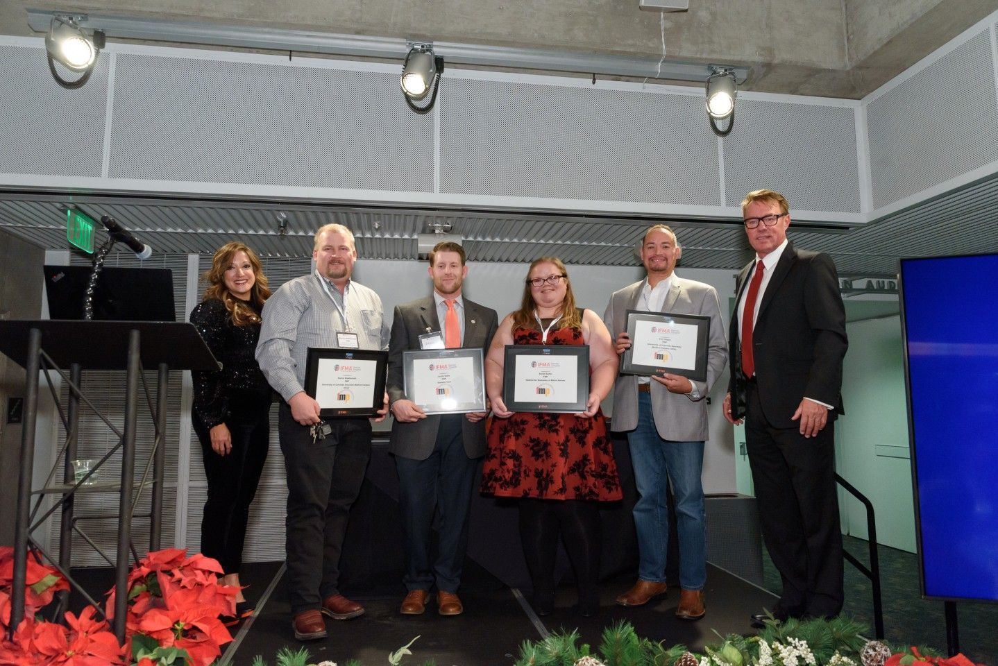 People on stage holding framed certificates, likely award recipients, in front of a blue screen and backdrop.