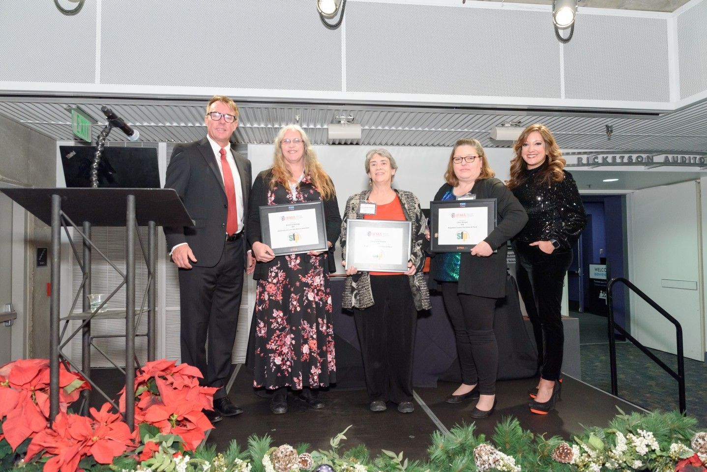 Five people on a stage holding certificates. A man in a suit stands by a podium. Others smile, with a festive backdrop.