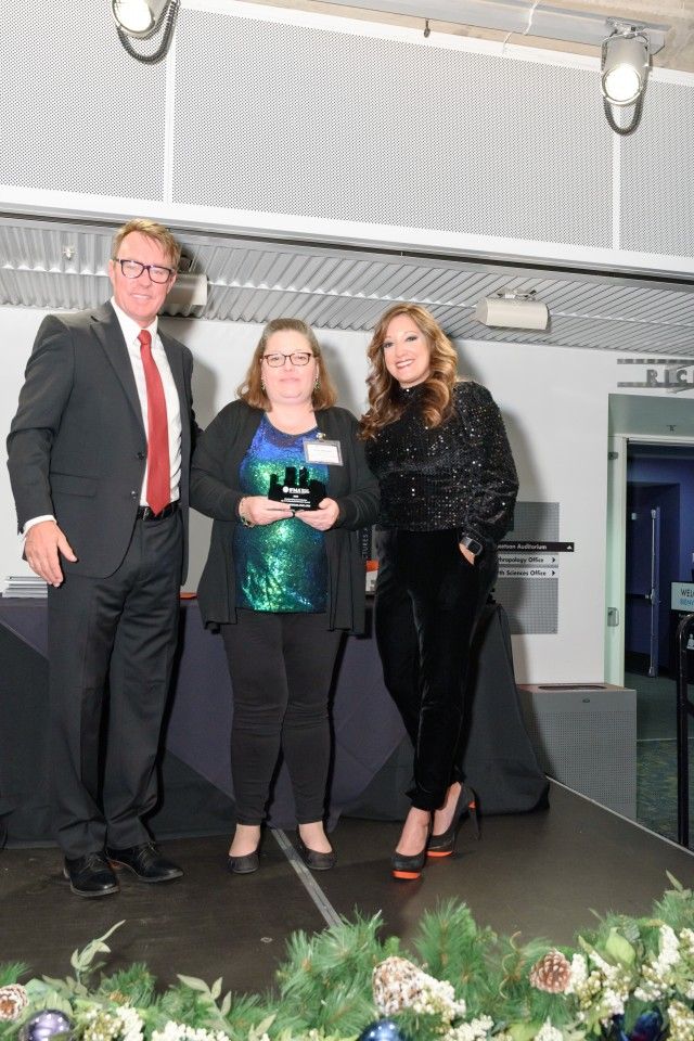 Three people on a stage: a man in a suit, a woman holding an award, and another woman smiling.