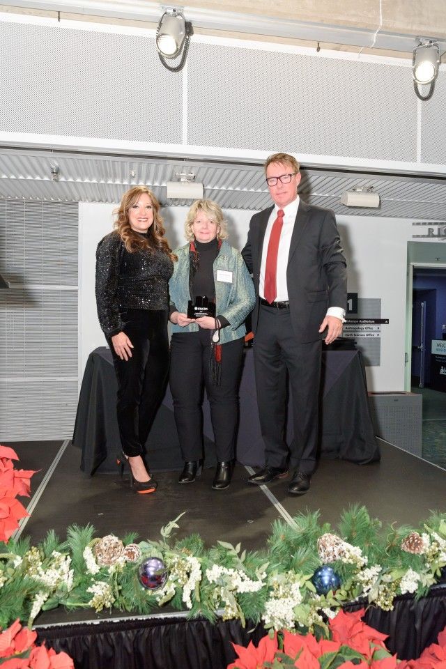 Three people on a stage with an award. A woman in the middle holds it. Decorations include poinsettias and greenery.