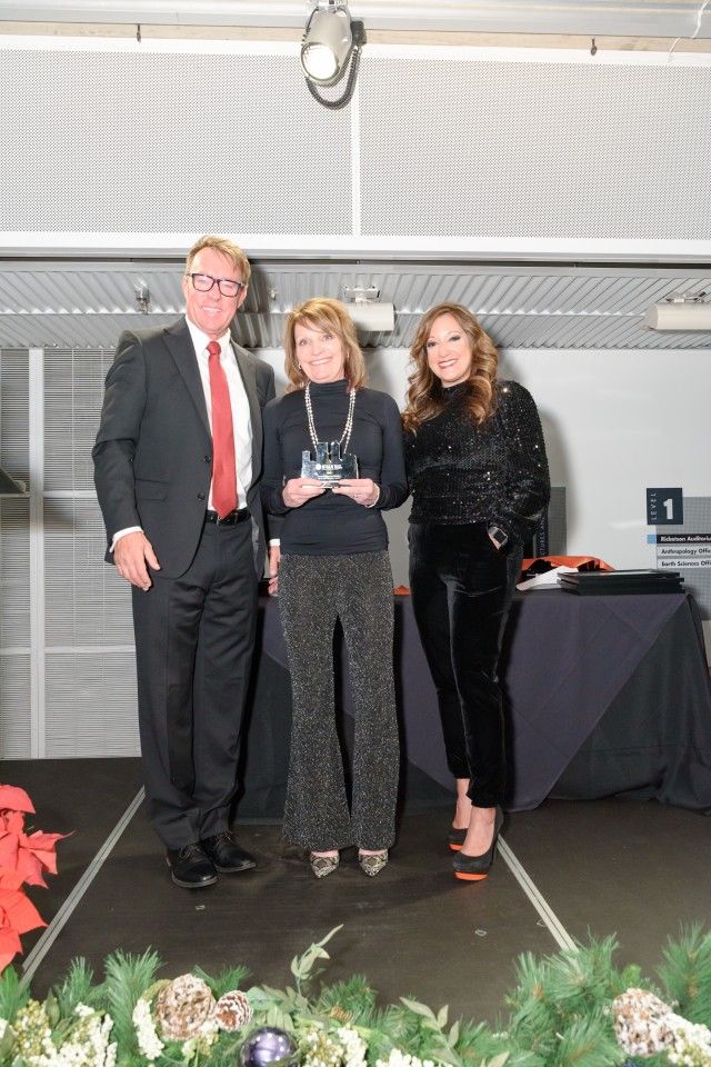 Three people on stage, one holding an award. Man in suit, two women in semi-formal wear, indoor event.