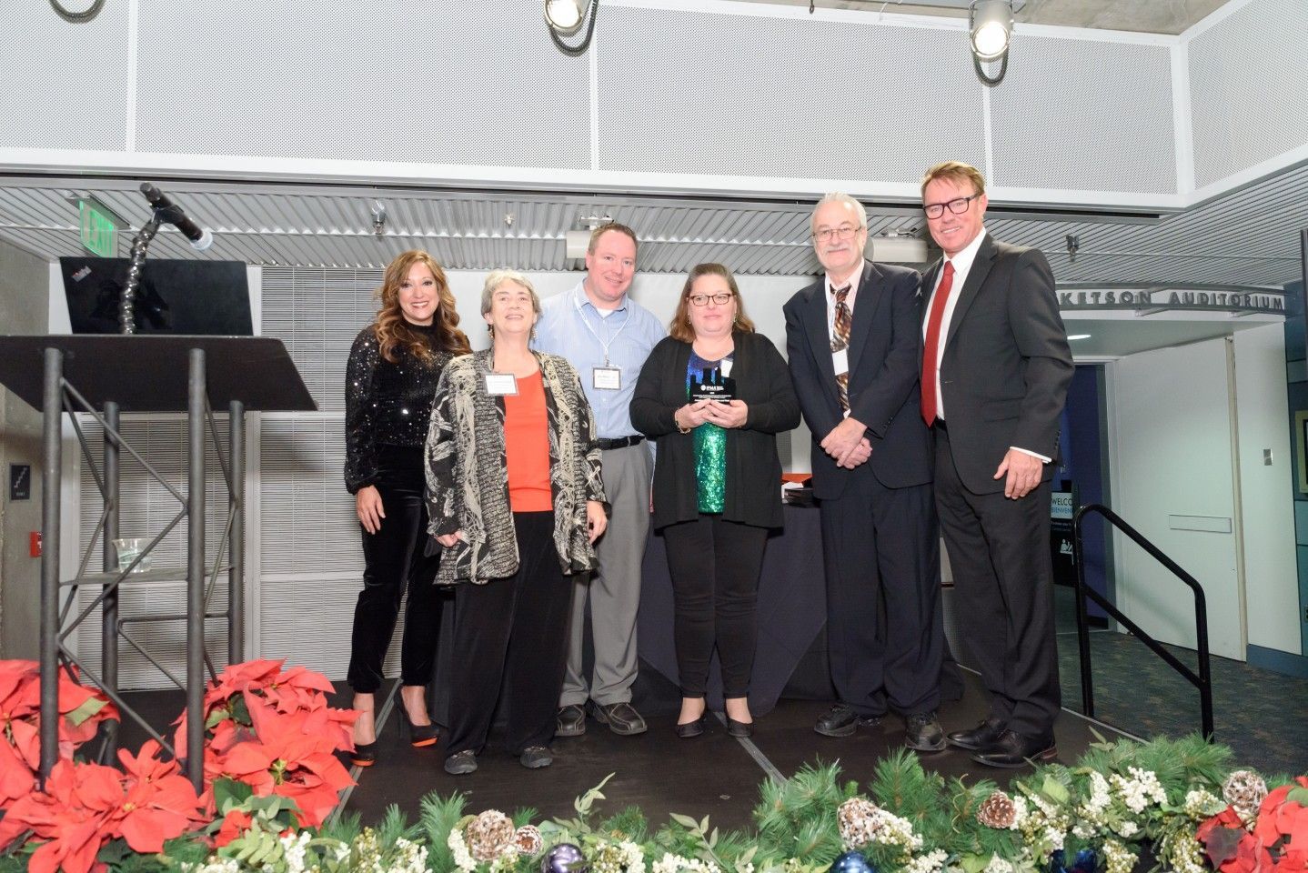 Group of people on stage with an award, red and green floral decorations.