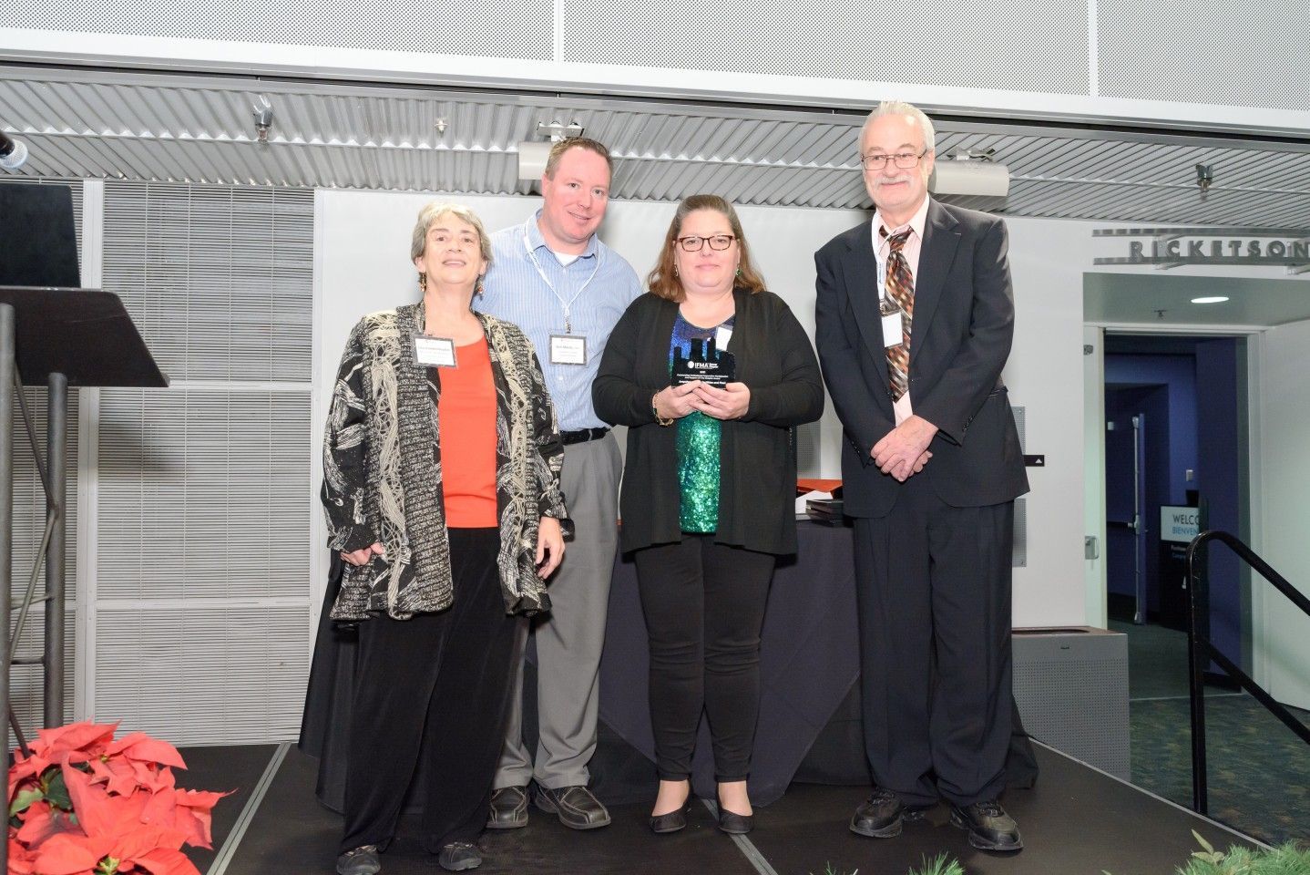 Four people on a stage holding an award. Two women, two men, with a silver and black backdrop.