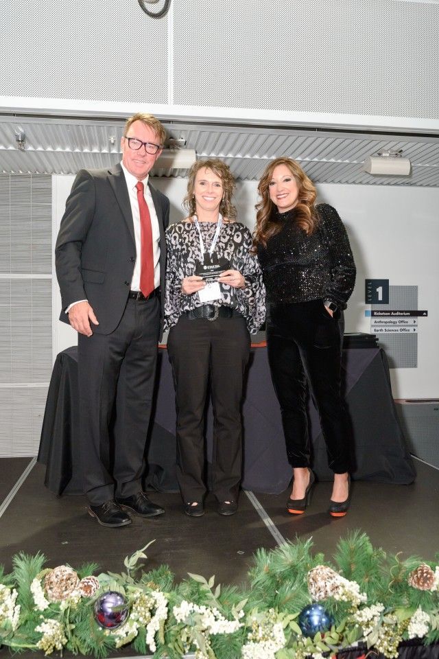 Three people on a stage: a man in a suit, a woman holding an award, and a woman in a sparkly top.