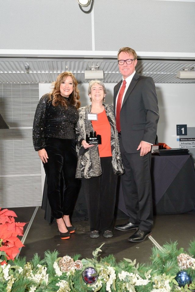 Three people on a stage; a woman holds a trophy. Festive decor.
