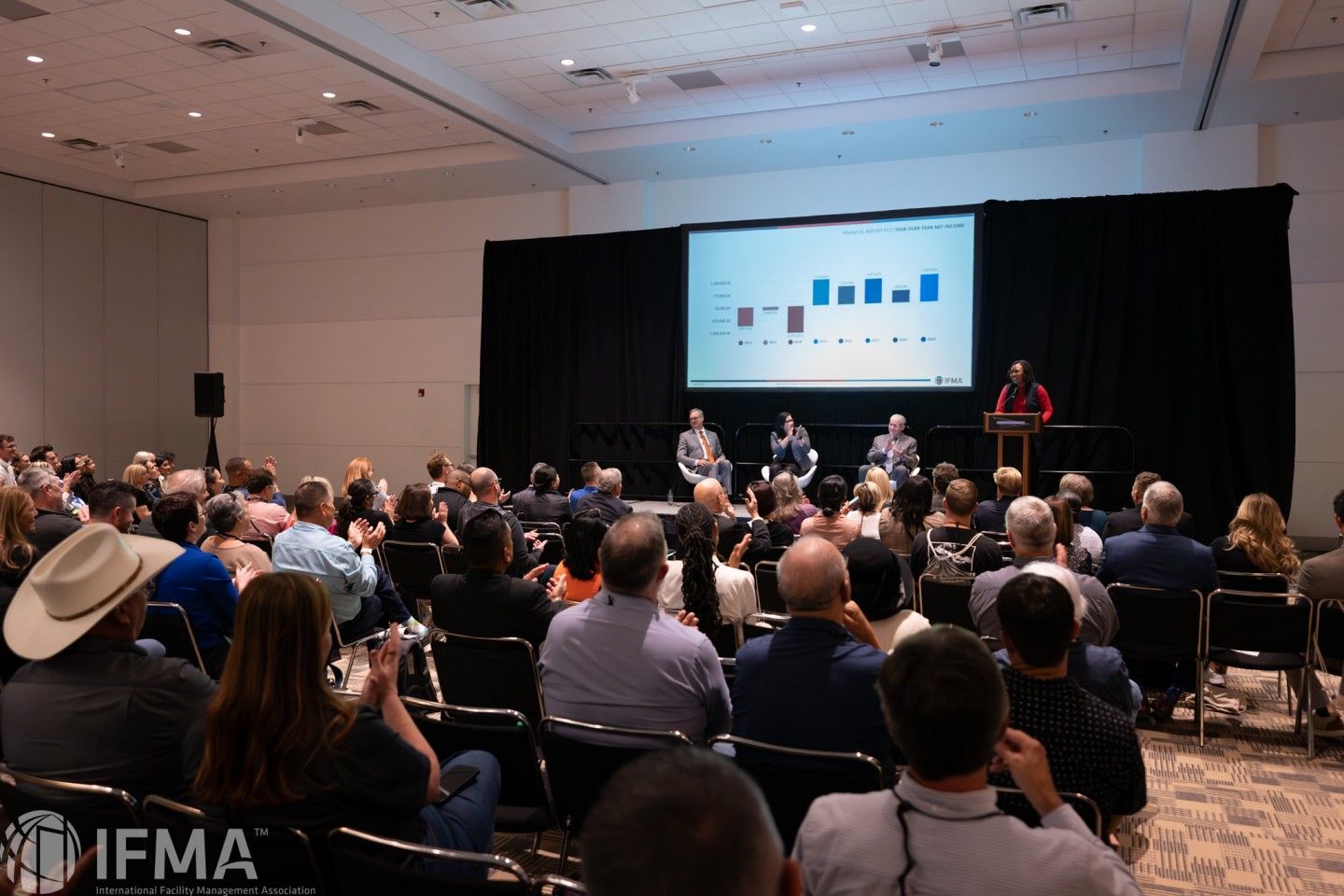 Audience in a conference room listening to a presentation on a stage with a graph displayed.