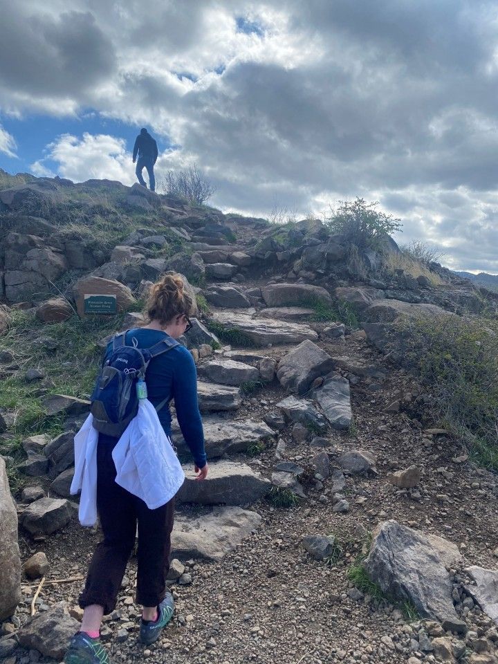 Person hiking up stone steps on a hillside trail. Another person is at the top under a cloudy sky.