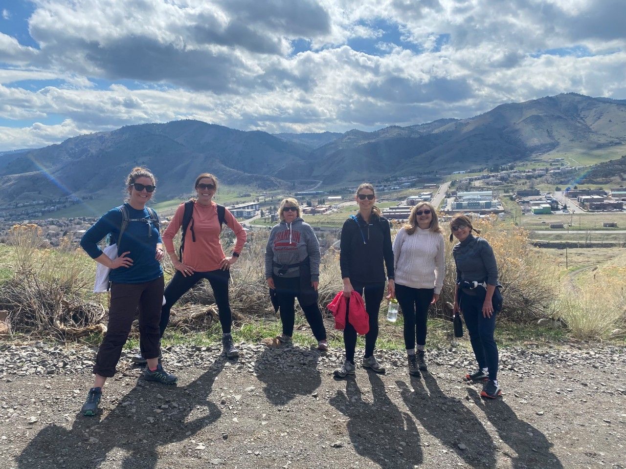 Six people stand on a hillside overlooking a town. Cloudy sky, mountains in the background.