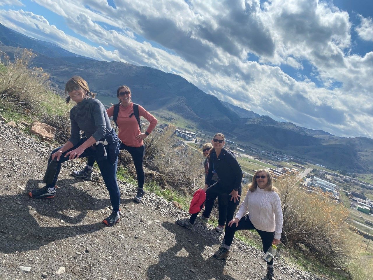 Four people on a hiking trail overlooking a valley. Cloudy sky, dirt path, and mountains in the background.