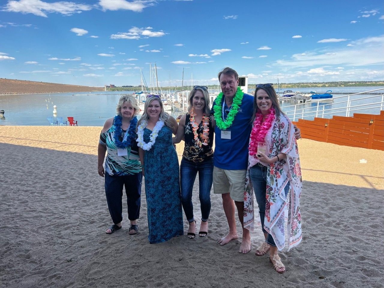 Five people with leis on a beach near a marina, smiling. Blue sky and water in the background.