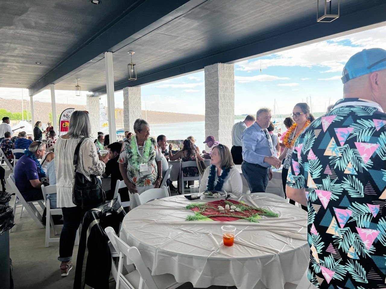 People at an outdoor event under a covered area, interacting, tables set with decorations, water in background.