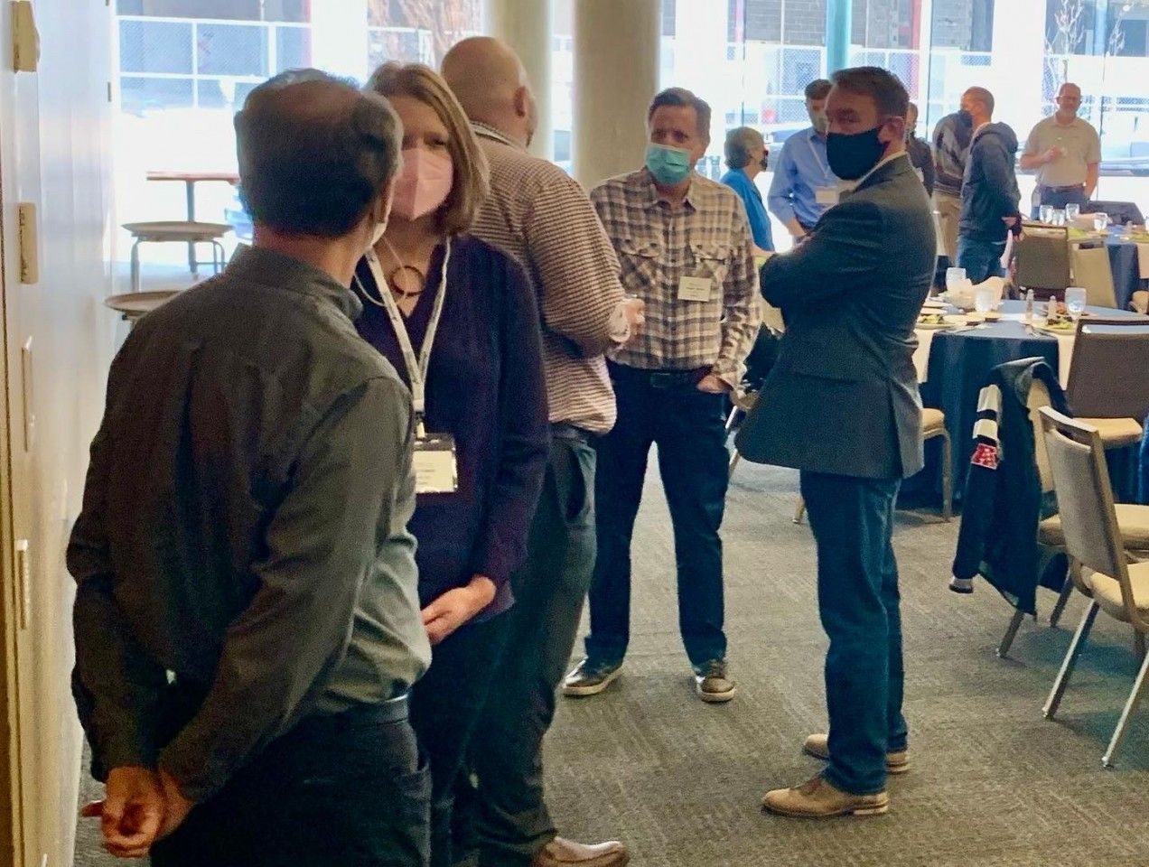 People in a conference room, wearing masks, conversing. Some are standing with name tags, others looking on.